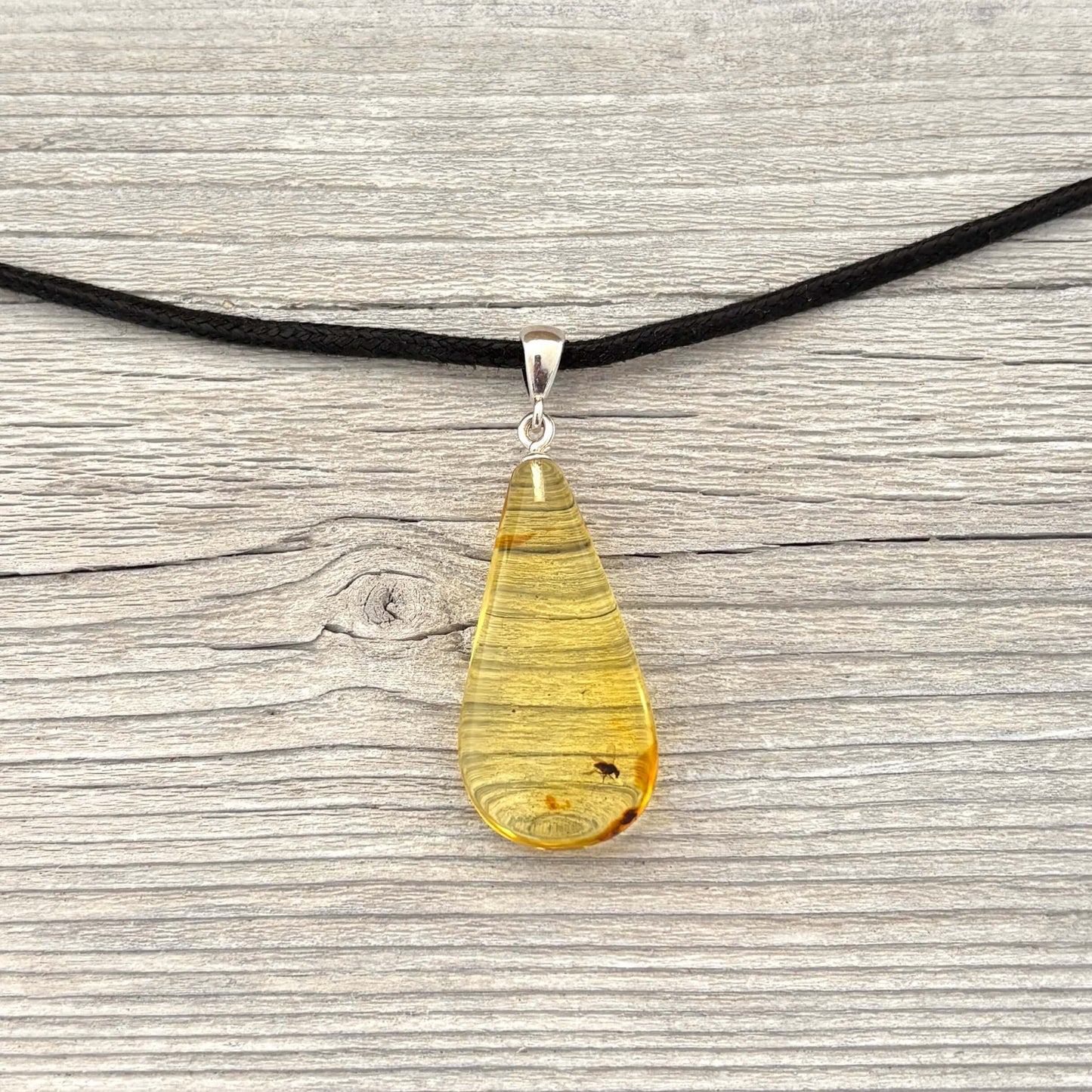 Teardrop-shaped amber pendant with insect on a black cord against a wooden background